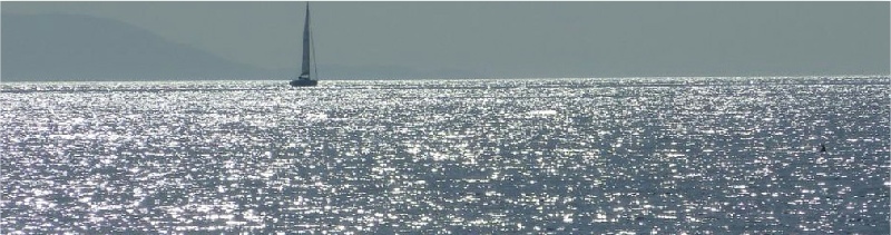 Segelboot im silbrig glitzernden Meer, links leicht verschwommen ein Hügel der Küste, fällt in Richtung Boot hin ab - Andreas Bertram-Weiss ǀ Mehr-Blick - Supervision- einen anderen Blick werfen 