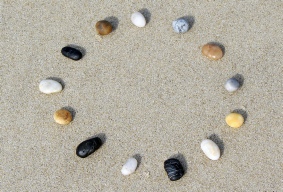Steinkreis aus dreizehn verschiedenfarbigen Steinen (weiss, braun, schwarz, blau, gelb) am Sandstrand aus der Vogelperspektive, das Licht der Sonne scheint von links hinter dem Betrachter - Mehr-Blick-Supervisionǀ Thurgau, Ostschweiz, Winterthur, St. Gallen und Schaffhausen